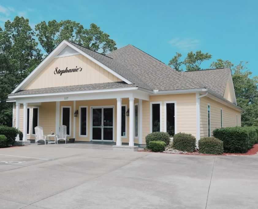 Yellow building with sign "Stephanie’s," porch chairs, and surrounded by greenery.